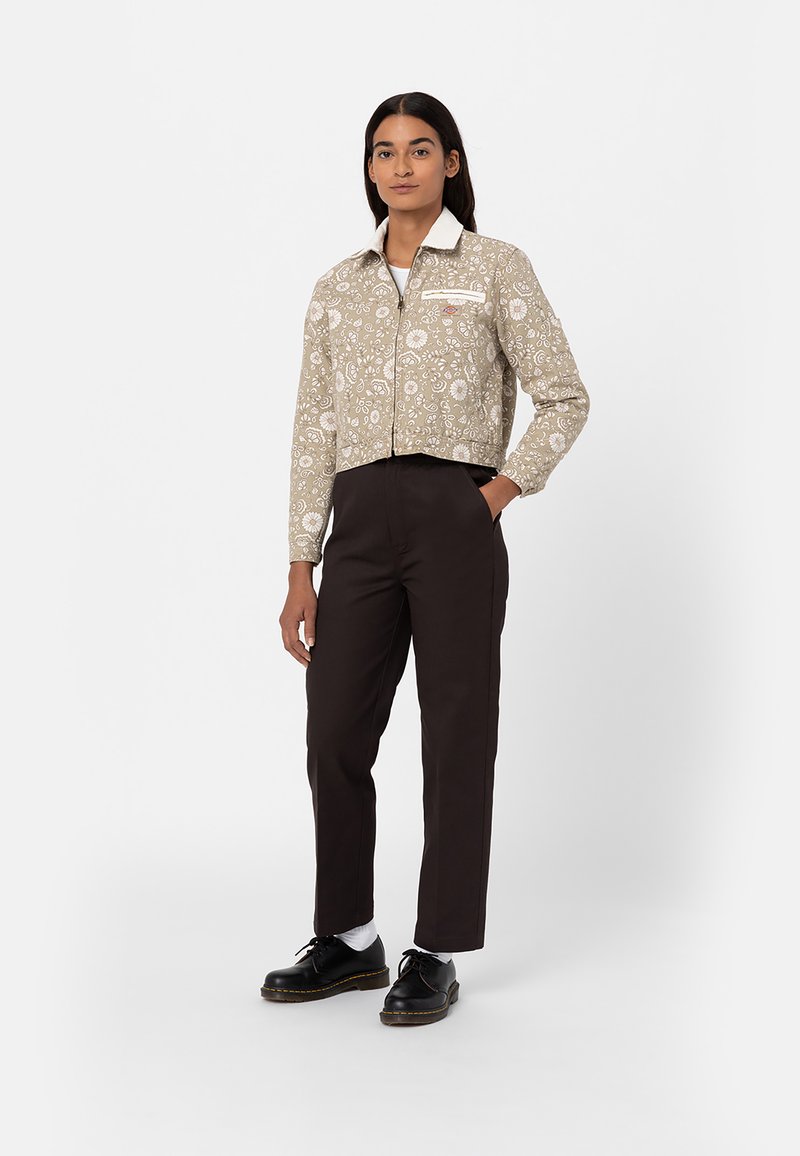 Chaqueta cremosa con cremallera y patrones florales blancos, cuello y bolsillo en el pecho, combinada con pantalones marrón oscuro y zapatos negros.