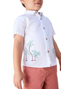 Chemise en lin blanc à manches courtes, ornée d'une broderie de palmier en vert et marron, fermeture par boutons en bois, associée à un short corail.