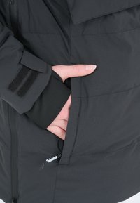 Svart isolerad jacka med dragkedja, sidofickor och kardborre-accenter vid ärmsluten. Materialet verkar slätt och matt.