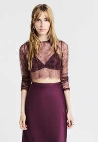 Top corto in pizzo bordeaux con lunghe maniche trasparenti sopra una gonna in satin abbinata. Presenta motivi floreali e un orlo leggermente frangiato.