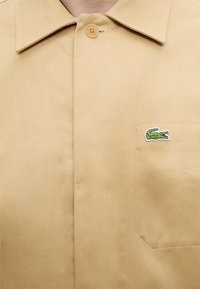Chemise beige avec col boutonné, poche poitrine et petit logo crocodile vert brodé sur la poche.