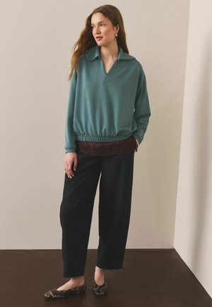 Žena stojící v tyrkysovém tričku s dlouhým rukávem a elastickým lemem, černých širokých kalhotách, hnědé spodní košili s ozdobnou krajkou a hadím vzorem na balerínkách, dívá se stranou.