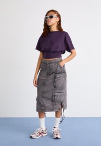 Lycra purppura, skirta pilka su kišenėmis, ir baltos kojinės iki kulniukų. Modelis dėvi saulės akinius ir sportinius batus su raudonais akcentais.