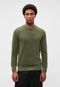 Alyvuogių žalios spalvos megztinis su apvalia iškirpte ir ilgomis rankovėmis. Turi tekstūruotą raštą ir subtilų logotipą krūtinės srityje.