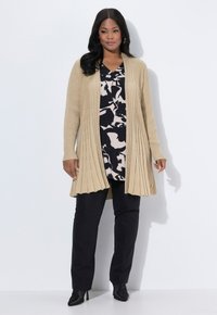 Mujer vistiendo un cárdigan de canalé beige sobre una blusa de patrón negro y blanco, pantalones negros y botas de tacón negras, de pie contra un fondo blanco.