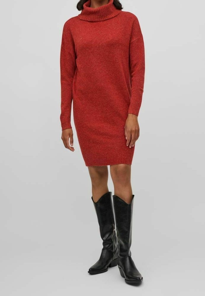 Femme portant une robe-pull en maille rouge à col roulé associée à des bottes en cuir noires montant jusqu'aux genoux, debout devant un fond neutre.
