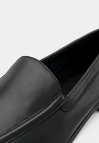Chaussure noire en cuir à enfiler, présentant une texture lisse, un design arrondi et des détails de couture subtils le long des bords.