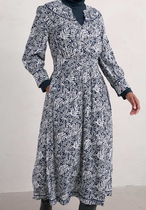 Woman wearing a navy blue and white floral midi dress layered over a black turtleneck, with hands partially in dress pockets.