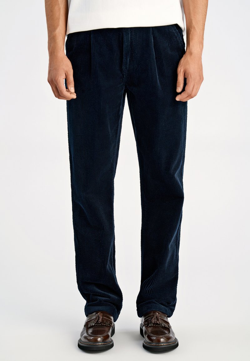 Marineblauwe corduroy broek met verticale geribbelde textuur, met een losse pasvorm en voorplooien, gecombineerd met bruine leren schoenen.