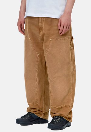 Persona che indossa pantaloni cargo marroni larghi con grandi tasche frontali e scarpe da trekking nere, in piedi davanti a uno sfondo semplice.