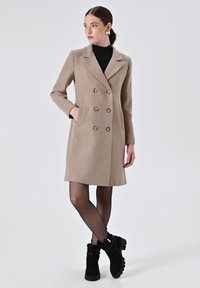 Cappotto beige a doppio petto realizzato in tessuto testurizzato, con bottoni dorati e tasche laterali, abbinato a stivali alla caviglia neri e collant.