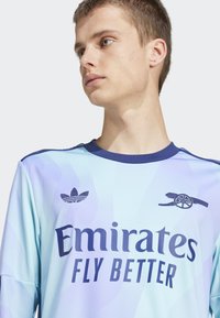 Lys blå fodboldtrøje med bølgemønster, der har mørkeblå detaljer, "Emirates Fly Better" tekst samt Adidas- og Arsenal-logoer på fronten.