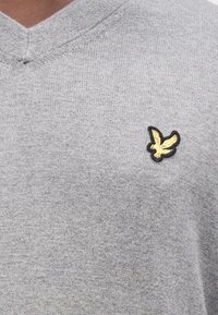 Šedý svetr s výstřihem do V z pletené látky s hladkou texturou. Na levé straně hrudi je malé černo-zlaté vyšívané logo ptáka.