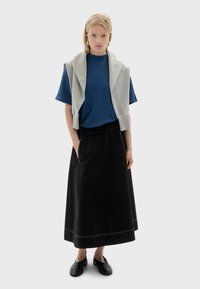 Frau in einem blauen kurzärmligen Oberteil, mit einem grauen Pullover über den Schultern, einem langen schwarzen Rock und schwarzen Slippers, die mit den Händen in den Taschen steht.