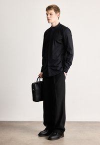 Fekete gombos ing mandzsettagallérral, széles fekete nadrággal és fekete bőr cipővel párosítva. Egy fekete téglalap alakú táskát tart.