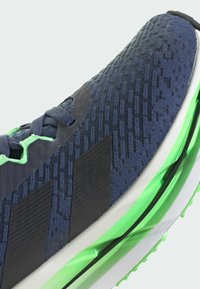 Close-up of a navy blue athletic shoe with black stripes, green accents, textured knit upper, and white and green cushioned sole.