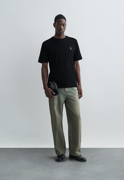 Joven con una camiseta negra, jeans holgados deslavados, zapatos negros, sosteniendo un bolso de mano negro con textura, de pie sobre un suelo gris frente a una pared lisa.