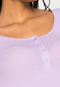 Lila ribbad topp med rundad halsringning och fem metallknappar i mitten. Mjuk textur med subtila horisontella ribbor.