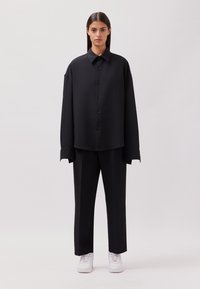Schwarzes, übergroßes Button-Up-Hemd mit langen Ärmeln, kombiniert mit passenden Hosen und weißen Sneakers. Glattes Material und strukturiertes Design.
