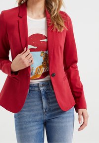 Röd figursydd kavaj, lätt stretchtyg, enkel knappstängning, spetsiga slag, buren över en grafisk t-shirt, tillsammans med blå jeans.