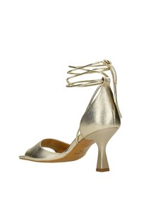 Metalické zlaté sandály na vysokém podpatku s designem peep-toe, kotníkovými pásky a štíhlým jehlovým podpatkem. Hladký povrch s minimálním zdobením.