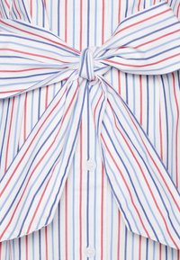 Koszula zapinana na guziki z pionowymi czerwonymi i niebieskimi paskami na białym tle, z wiązanym akcentem z tkaniny w talii i przezroczystymi guzikami.