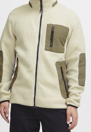 Personne portant une veste polaire crème avec des poches zippées vert olive sur la poitrine, les côtés et les coudes, et une fermeture éclair noire à l'avant.