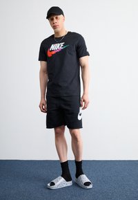 T-shirt preta da Nike com logótipo colorido, calções pretos com logótipo branco, meias pretas e chinelos brancos com padrão sobre um chão carpete azul.