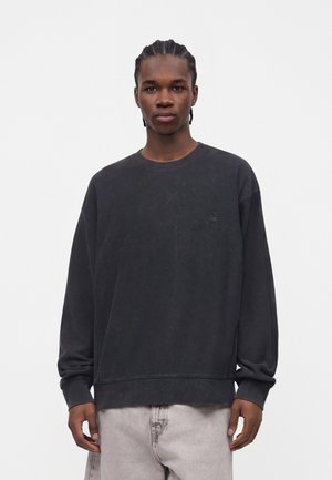 Jeune homme aux cheveux tressés portant une chemise noire ample à manches longues et un pantalon gris clair, debout sur un fond blanc uni.
