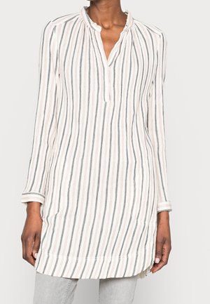 Tunique blanche à manches longues avec des rayures verticales noires et roses, encolure fendue et manches avec poignets, portée par une personne portant un pantalon gris clair.