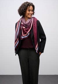 Sciarpa colorata a motivo triangolare con tonalità di bordeaux, rosa e blu, sovrapposta a una giacca e a un maglione scuri, creando profondità visiva.
