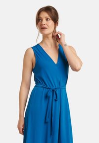 Blue sleeveless dress in a smooth fabric, featuring a V-neckline and a tied waist. The design offers a flowing silhouette and is mid-length.