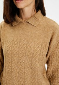 Suéter de punto marrón claro con un patrón de espiga texturizado, cuello doblado y mangas largas, hecho de un material suave.