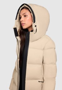 Woman wearing beige padded winter jacket with large hood and black zipper details, facing left against a plain gray background.