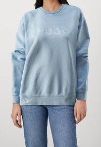 Sudadera azul claro con cuello redondo, corte oversized y puños acanalados. Presenta el logotipo bordado "HUGO" en el pecho. Textura suave.
