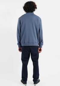 Personne aux cheveux bouclés portant un haut à manches longues bleu, un pantalon bleu foncé et des baskets noires, debout de dos sur un fond blanc.