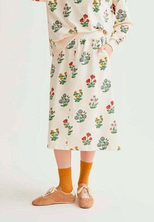 Persona con una falda floral color crema y un top a juego, calcetines naranjas y zapatillas marrones con textura, de pie con una mano en el bolsillo.