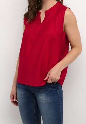 Vrouw draagt mouwloze rode blouse met kleine plooien en blauwe spijkerbroek, hand rustend nabij zak tegen een effen achtergrond.