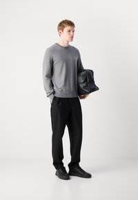 Junger Mann in einem grauen Pullover, schwarzen Hosen und schwarzen Turnschuhen, der eine große schwarze Ledertasche vor einem weißen Hintergrund hält.
