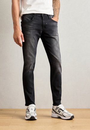 Jean slim fit en denim noir avec une finition délavée, design à cinq poches et détails de couture subtils, associés à des chaussures de sport blanches.