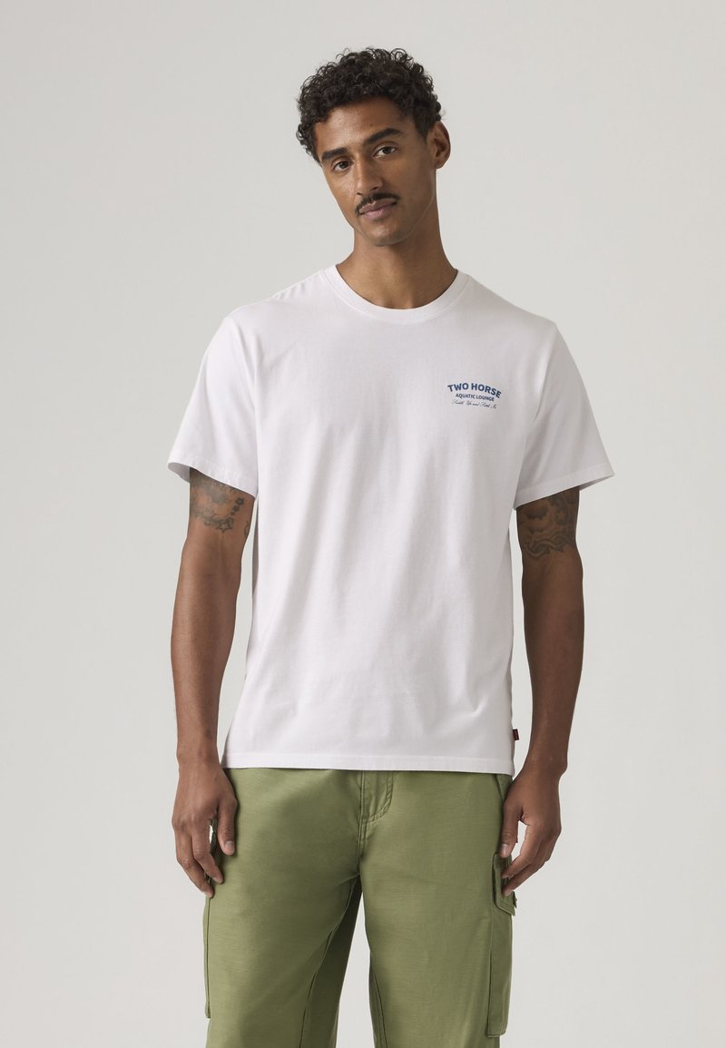 Mann mit Tätowierungen, der ein weißes T-Shirt mit dem Schriftzug "Two Horse Aquatic Lounge" und olivgrüne Cargohosen trägt, steht vor einem schlichten Hintergrund.