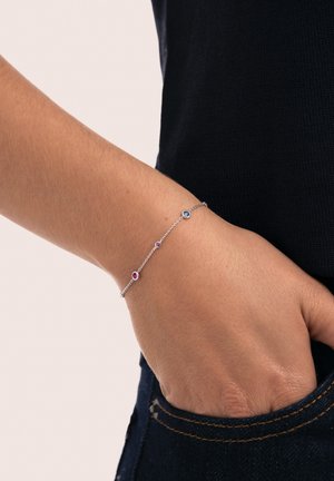 Pulsera de cadena de plata con tres pequeñas piedras de colores: azul, rosa y púrpura, colocadas en un diseño delicado. Mano descansando en un bolsillo de mezclilla.