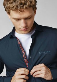 Tamsiai mėlynas marškinis su mygtukais, su "Red Bull Racing" logotipu, kontrastingomis raudonomis siūlėmis palei mygtukų eiles ir lygia tekstūra. Slim fit, šiuolaikinis dizainas.