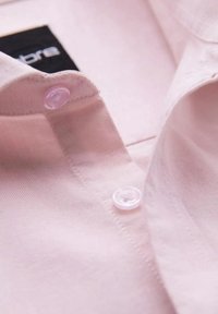 Chemise rose pâle à col avec coutures visibles, deux boutons translucides fermés et une étiquette de marque noire et blanche à l'intérieur du col.