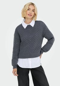 Blonde Frau, die einen grauen Strickpullover über einem weißen Button-Up-Hemd und schwarzer Hose trägt, posiert mit einer Hand auf der Hüfte.