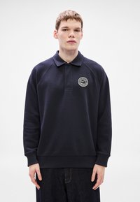 Μπλε σκούρο πόλο φούτερ με γιακά, μακριά ραγκλάν μανίκια και στρογγυλό έμπλαστρο λογότυπου στο στήθος. Το ύφασμα φαίνεται μαλακό και με υφή.