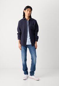Mørkeblå teksturert knappeløs skjorte med brystlomme, kombinert med lyseblå t-skjorte, blå jeans og hvite sneakers.