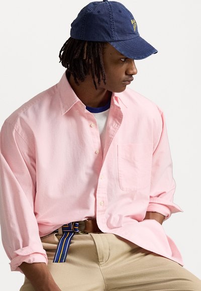 Chemise boutonnée rose clair à manches longues, avec une poche poitrine unique, portée par-dessus une chemise blanche, associée à un pantalon kaki et une casquette de baseball bleue.