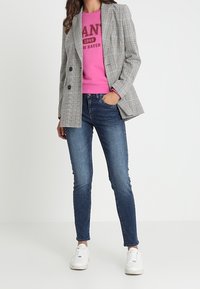 Blazer doppiopetto a quadri grigi sopra una felpa rosa, abbinato a jeans blu e sneakers bianche. Outfit casual e stratificato.