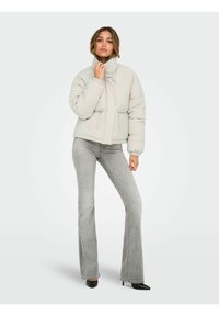 Chaqueta de plumas gris claro con cuello alto, bolsillos con cremallera y puños elásticos, combinada con vaqueros acampanados grises y tacones negros.